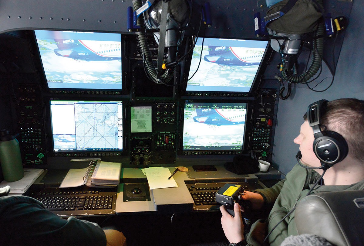 An operator wearing headphones uses a joystick at a mission control console inside an aircraft, monitoring multiple screens that display maps and a live video feed of an MSRC airplane.