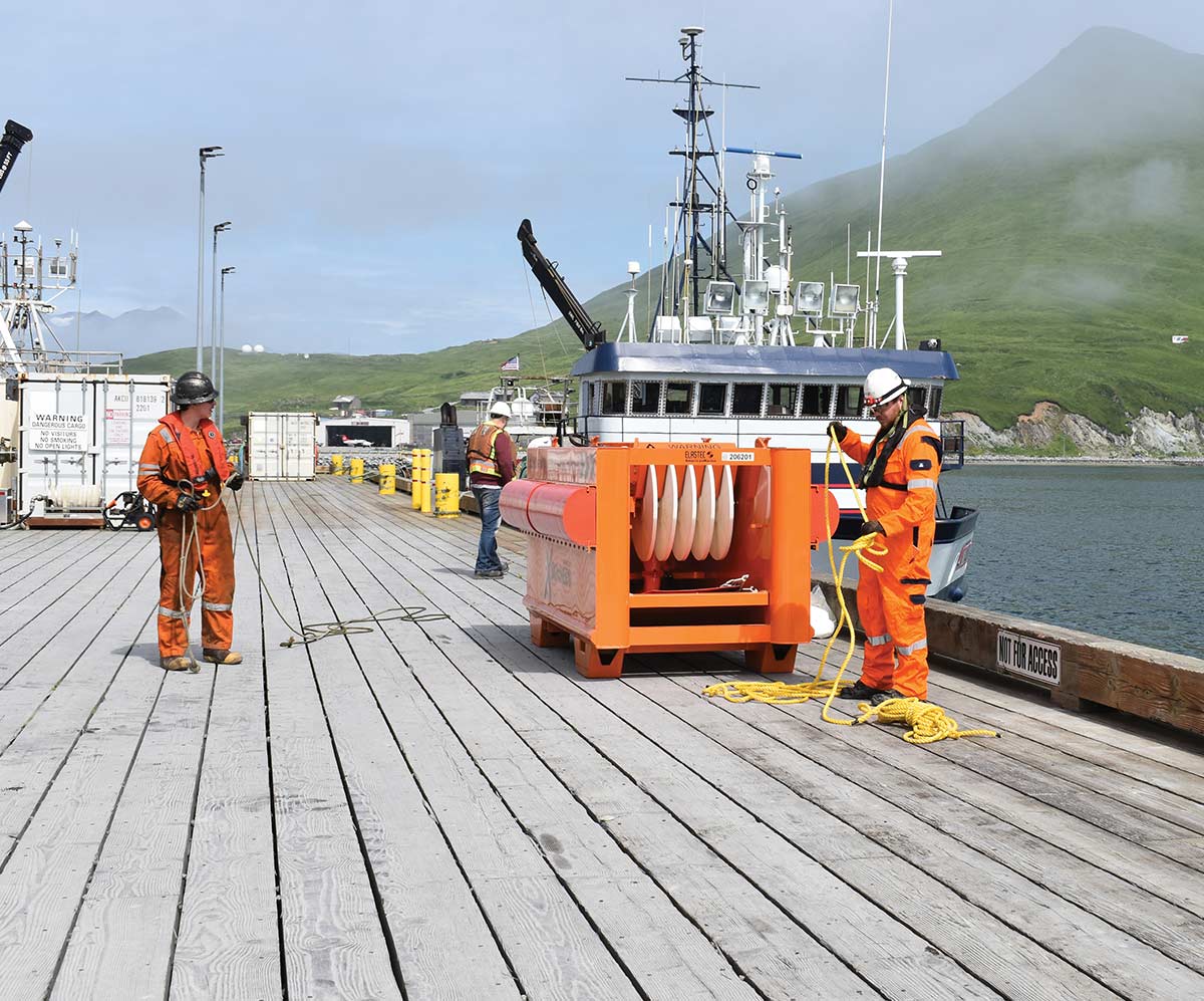 Two workers in orange safety gear handle yellow ropes on a wooden dock next to a large orange equipment unit, with a vessel and foggy green hills in the background.