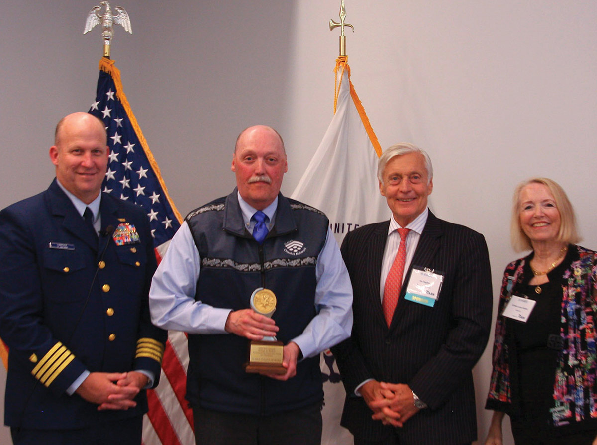 A Coast Guard officer, a man holding a circular award, a man in a suit, and a woman smile and pose together in front of the U.S. flag and a white organizational flag.