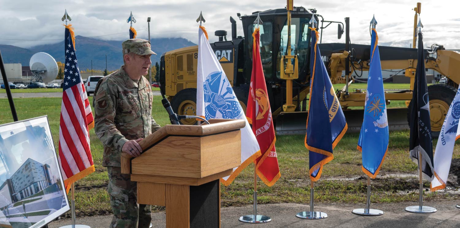 A military officer speaks at a wooden podium outdoors, flanked by a row of service flags with a yellow Caterpillar excavator behind him.