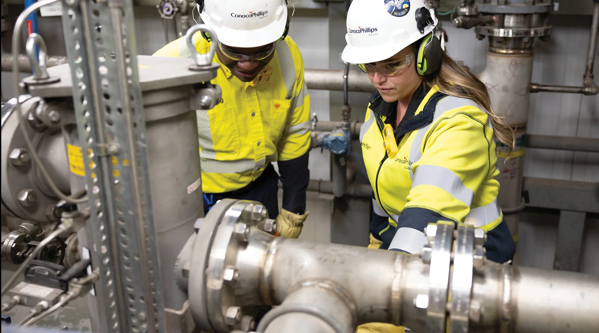Two workers in hard hats, hearing protection, and high-visibility clothing inspect piping and equipment at an industrial facility.