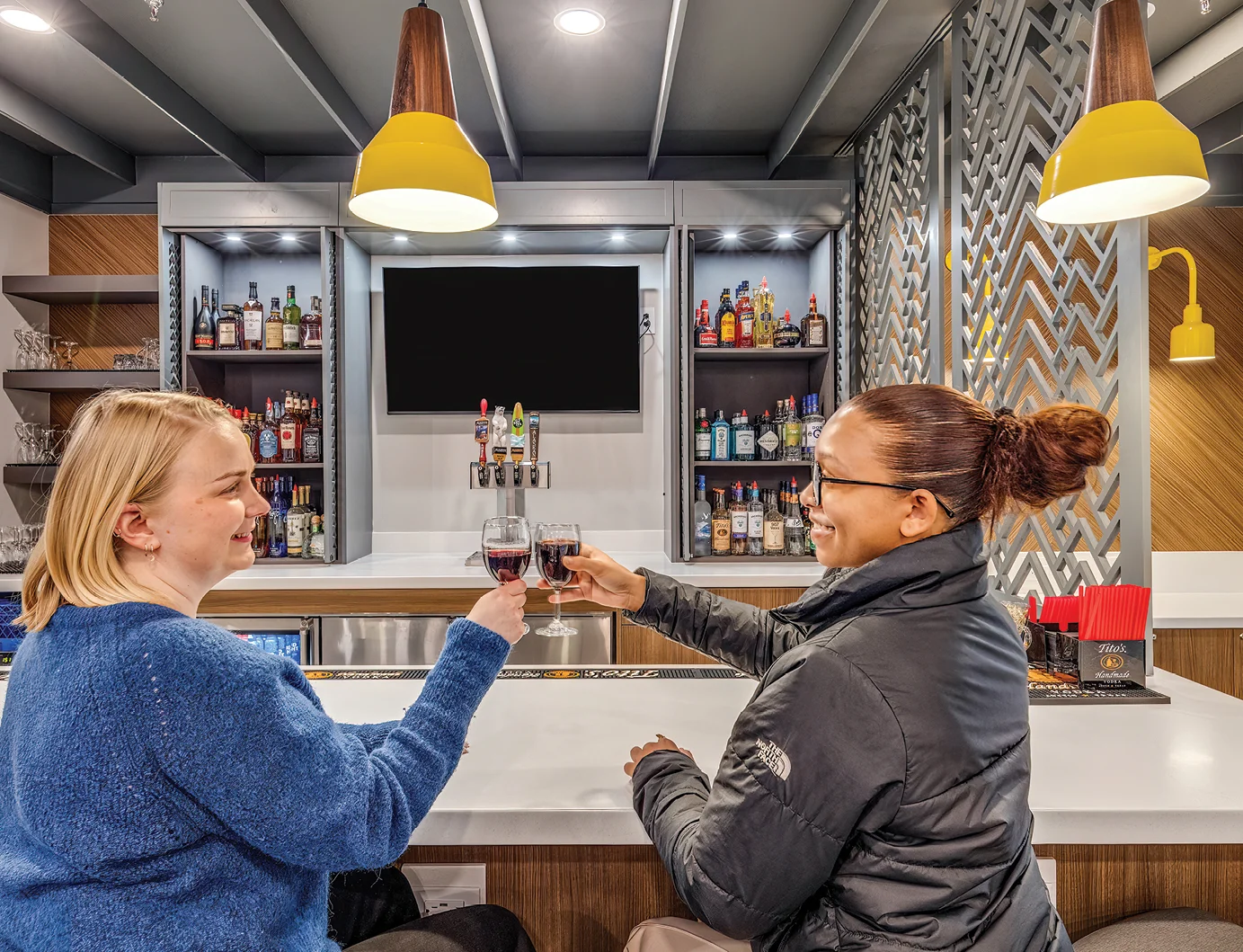 Two women seated at a hotel bar smile and raise glasses of red wine in a toast.