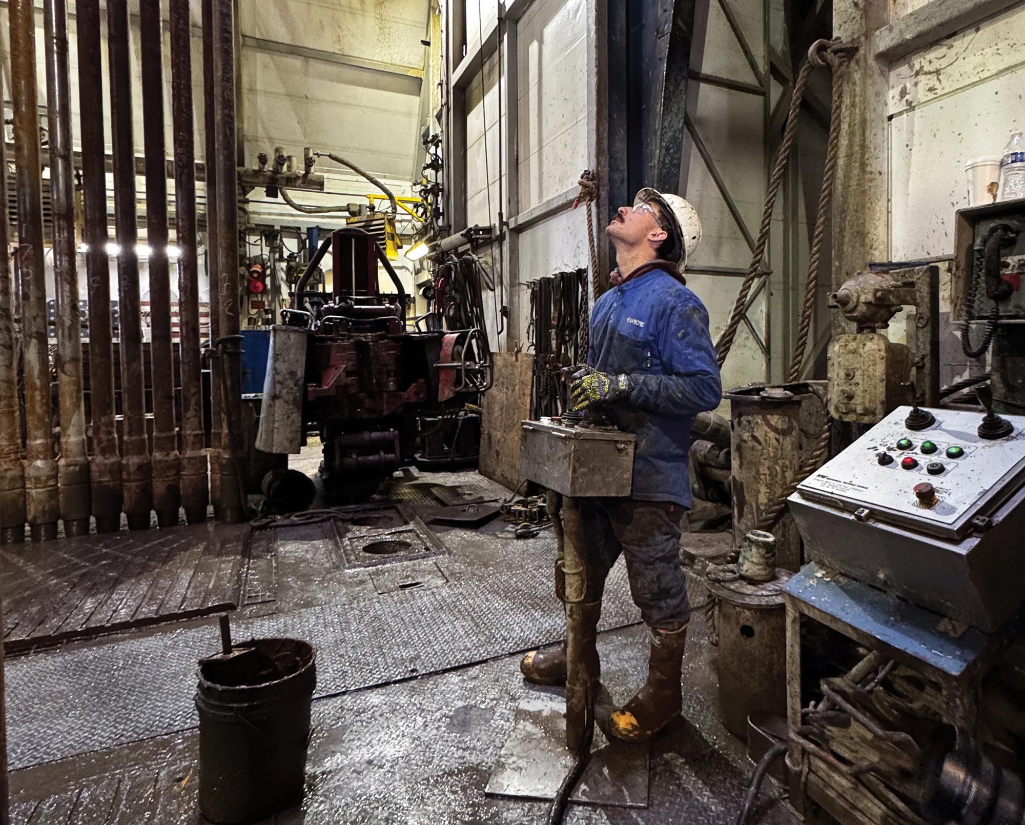 Worker in protective gear operates equipment beside rows of drilling pipe inside an industrial facility.