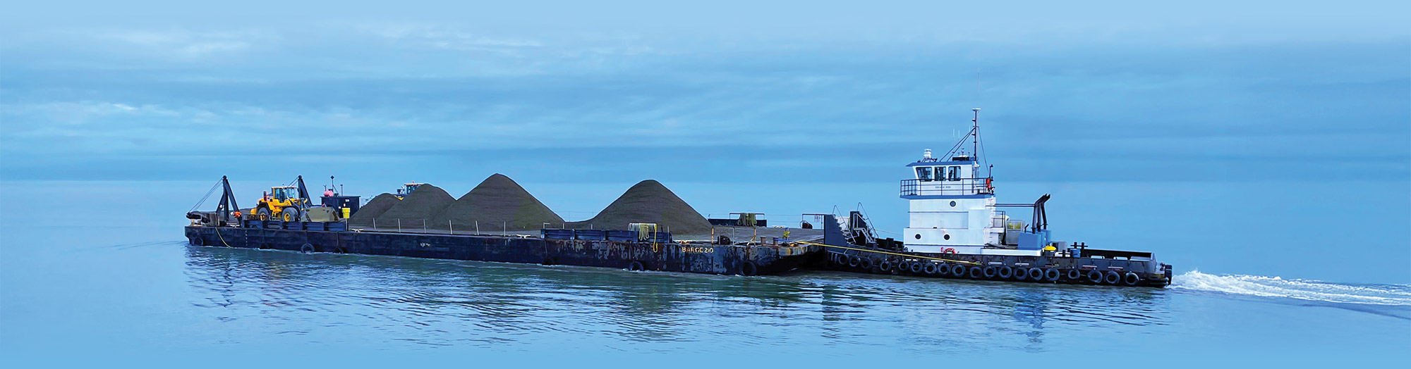 A white and blue tugboat pushes a large flat barge loaded with mounds of gravel and a yellow front loader across calm water under a cloudy sky.