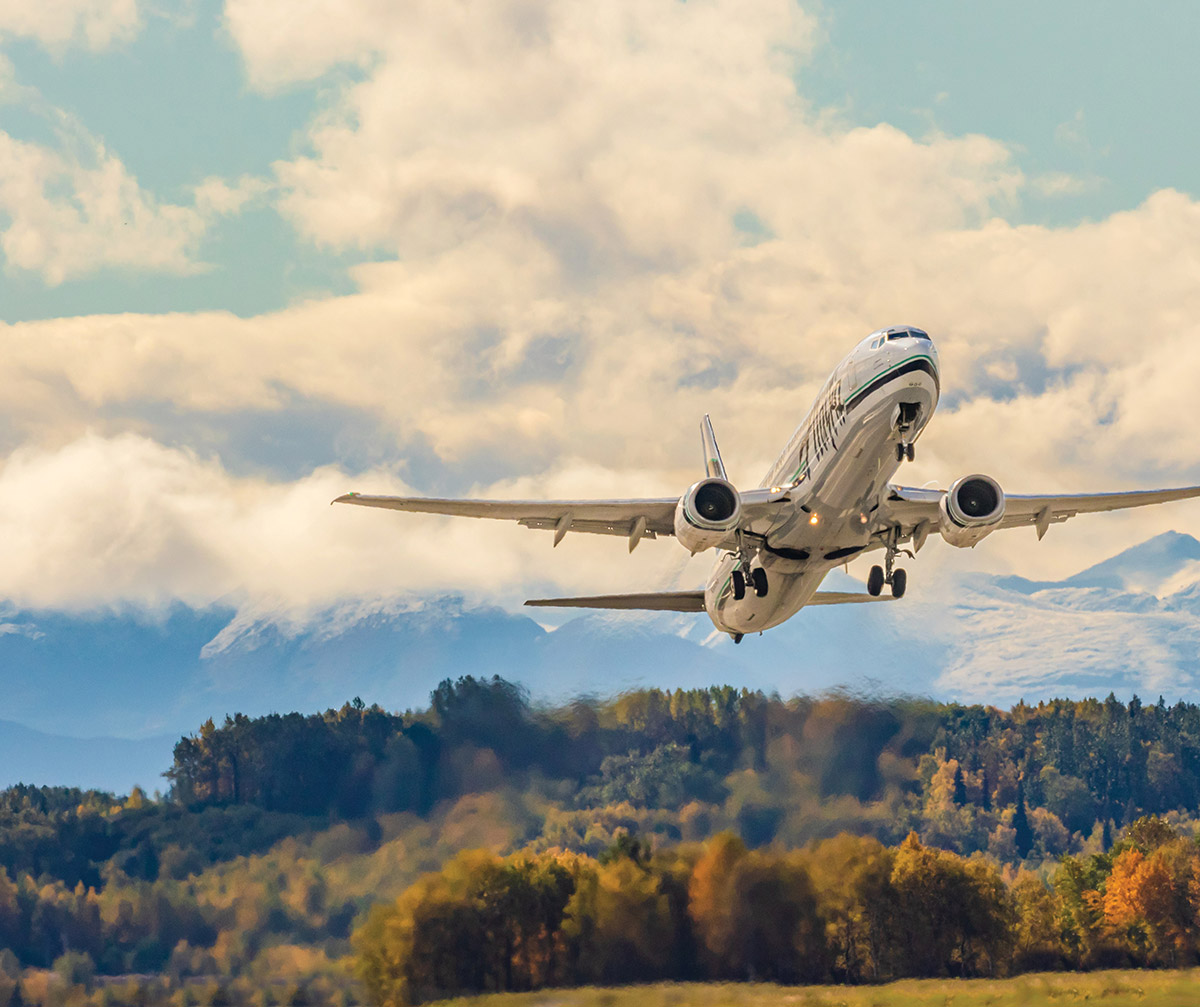 An airplane takes off over a colorful autumn forest with snow-capped mountains in the background.