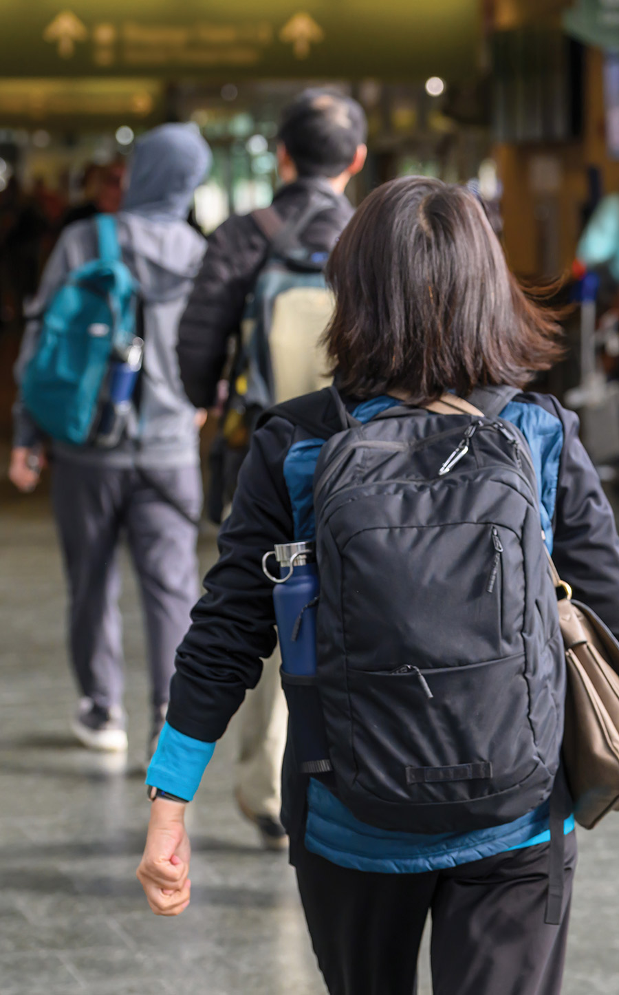 Back view of travelers wearing backpacks walking through a busy airport terminal corridor.