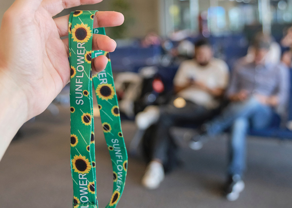 A hand holds up a green lanyard printed with sunflowers in front of blurred passengers.