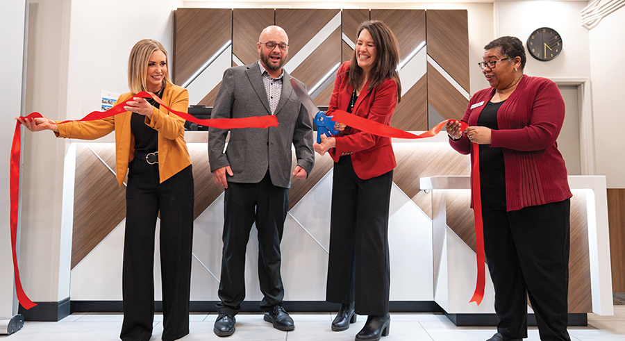 Four people celebrate a grand opening by cutting a large red ribbon with scissors.