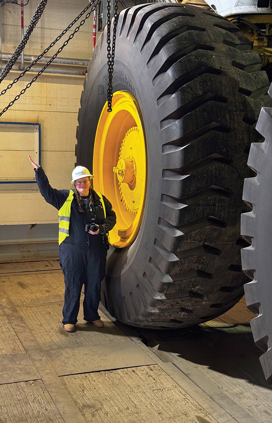 A worker in safety gear waves while standing next to a massive industrial tire.