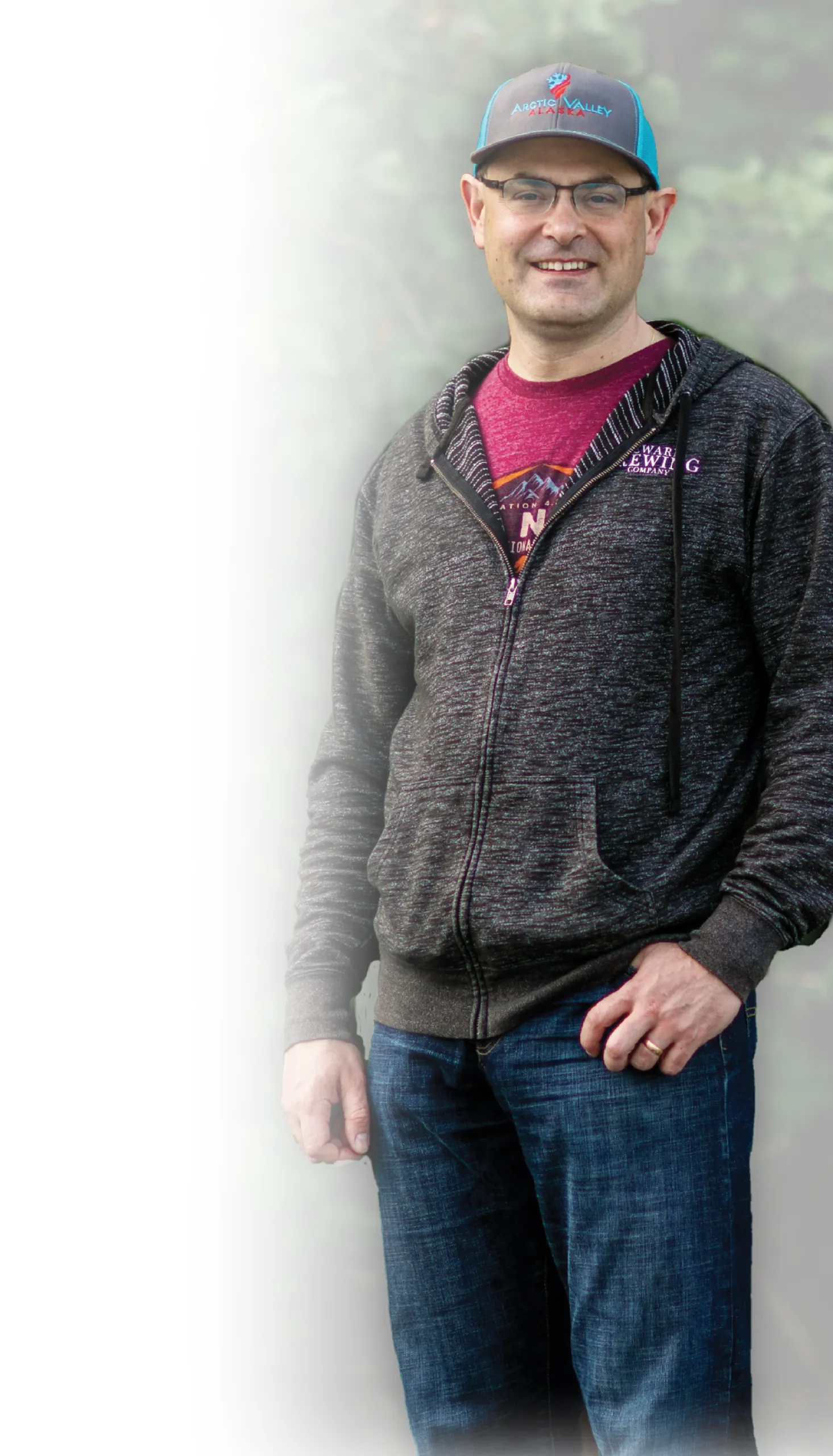 Portrait of a smiling man wearing glasses, a dark hoodie, and an Arctic Valley Alaska cap.