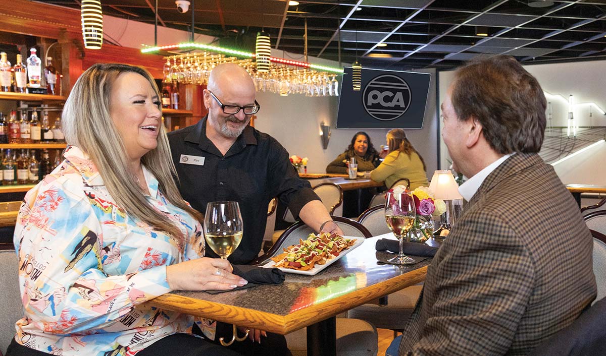 A server serves a plate of nachos to a couple enjoying wine in a restaurant.