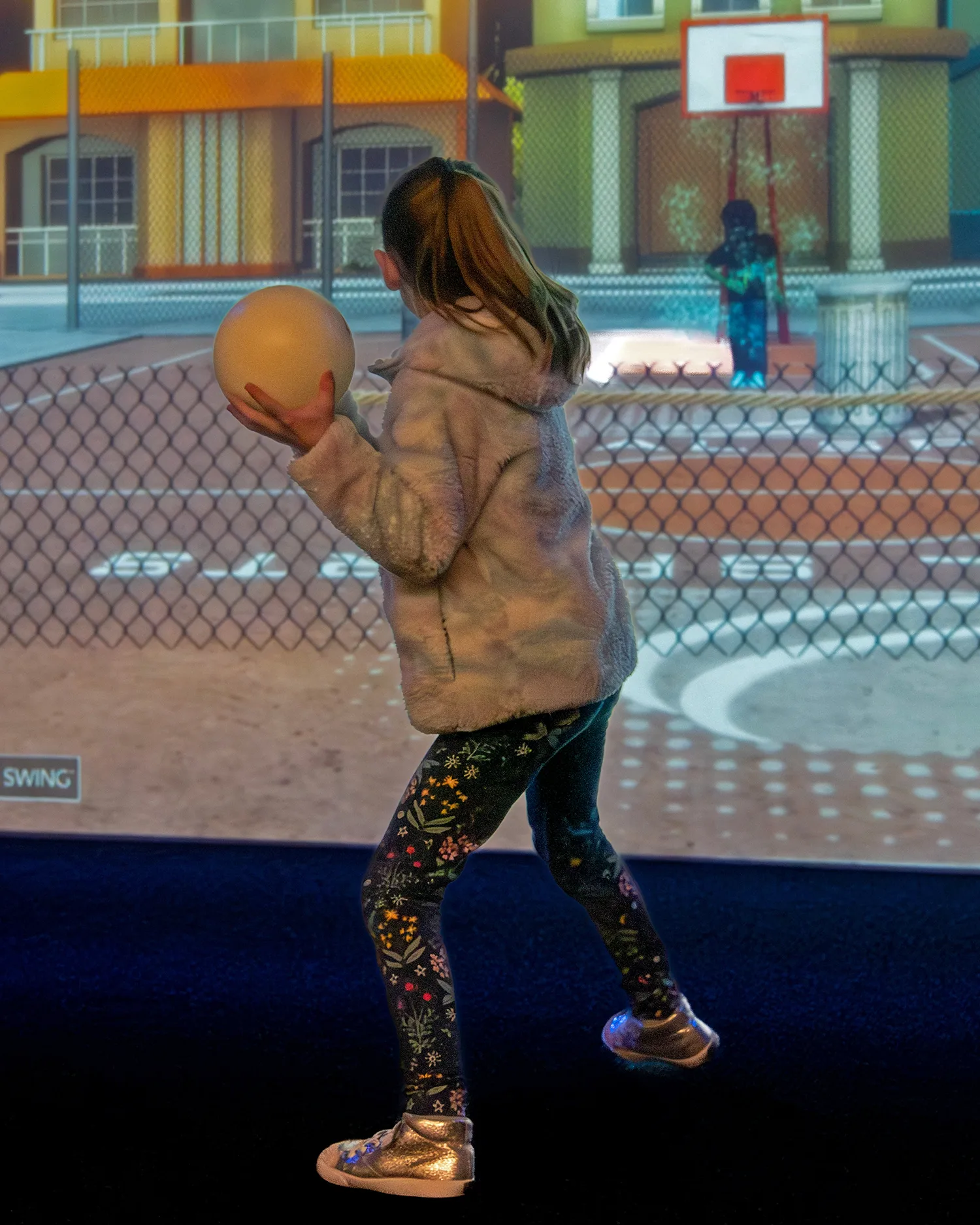 A child holding a ball beside a fenced indoor recreation court.