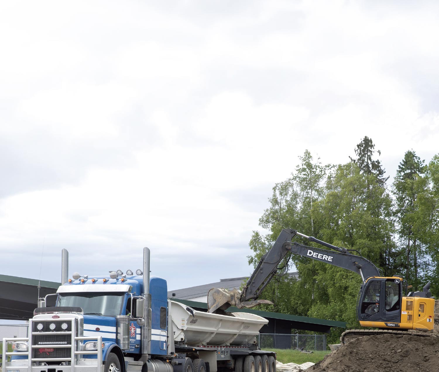 A yellow and black John Deere excavator scoops dirt into the bed of a large blue semi-truck at an outdoor job site.