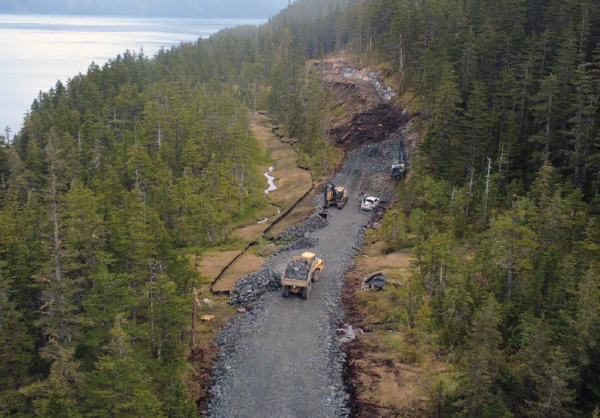 High-angle view of construction vehicles building a gravel road along a forested shoreline.