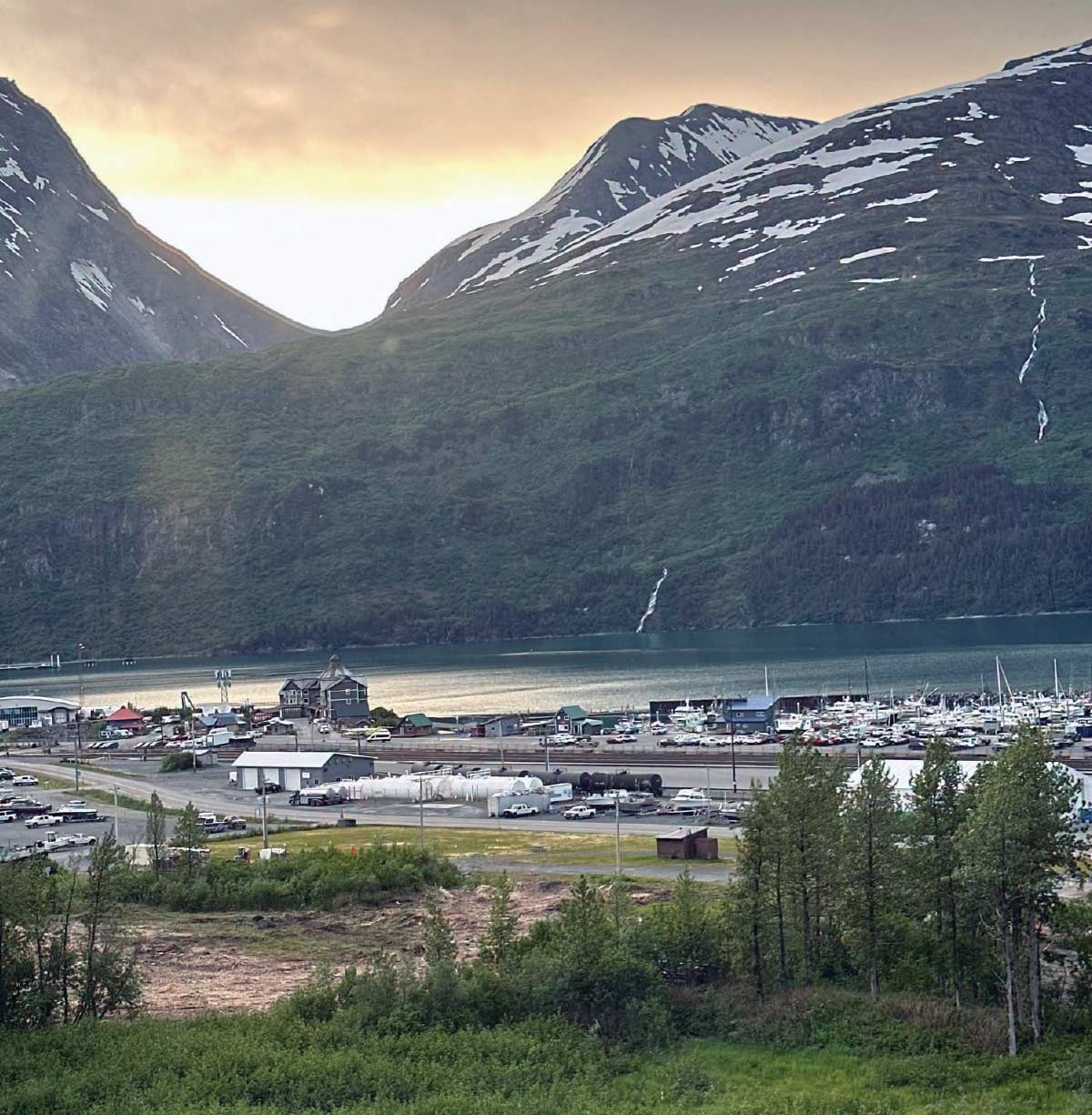 Scenic view of a coastal harbor filled with boats at the base of steep, snow-patched mountains.