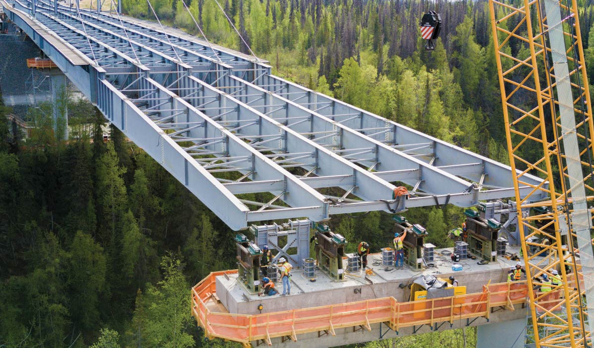 Construction crew working on steel girders during bridge assembly over a forest gorge with crane support.