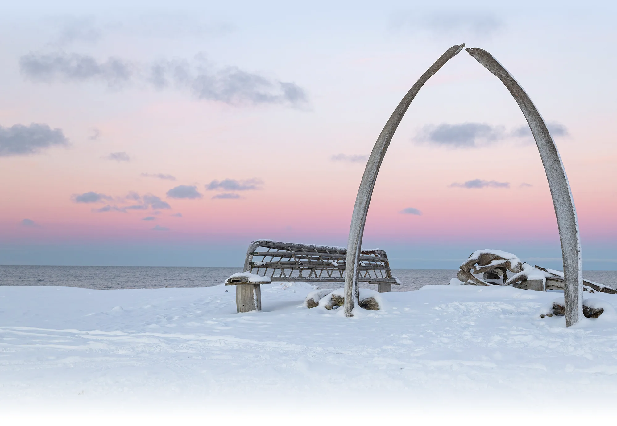 A tall whale jawbone arch and a wooden bench stand on a snow-covered shore, overlooking calm water under a soft pink and blue sunset sky.