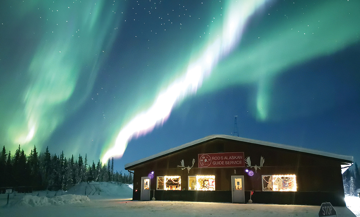 Vibrant green northern lights illuminate the starry night sky above a lit building with a "Rod's Alaskan Guide Service" sign in a snowy winter landscape.