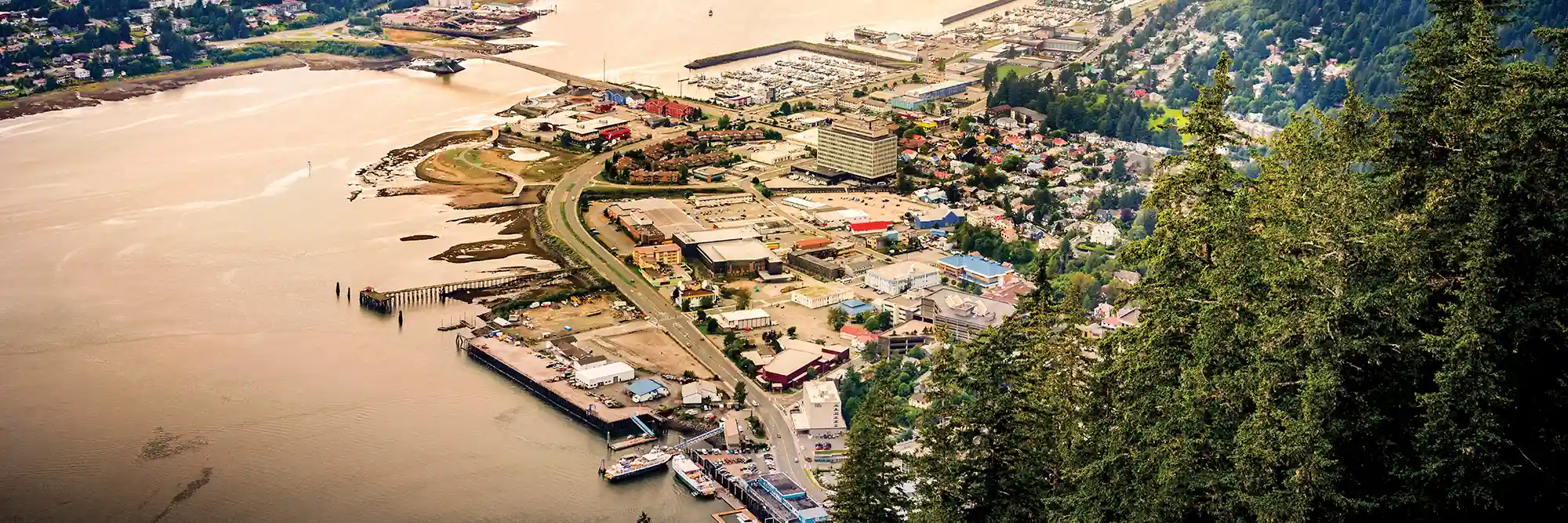 High-angle aerial view of a coastal town along a wide waterway, framed by evergreen trees, featuring harbor docks, boats, and waterfront buildings.