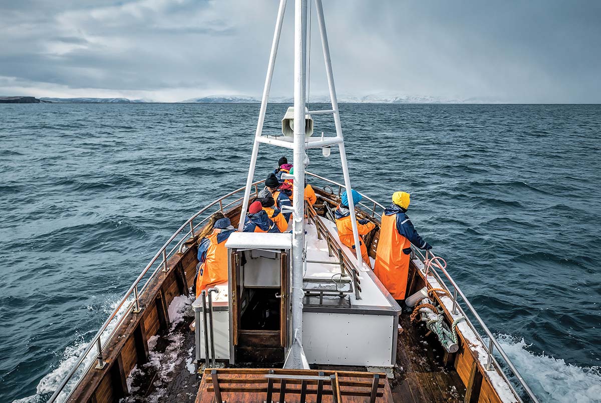 Passengers wearing bright orange rain gear gather at the bow of a wooden boat navigating choppy open waters, with distant snow-covered hills under an overcast sky.