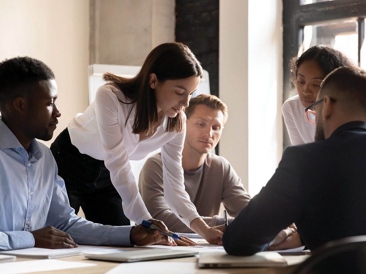 A diverse team of professionals collaborating around a table during an office meeting, with a woman leaning forward to review documents with her colleagues.