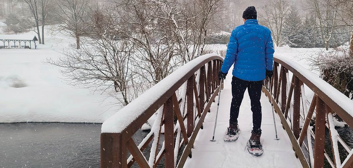 A person in a blue jacket snowshoeing across a snow-covered footbridge using trekking poles during a snowfall.