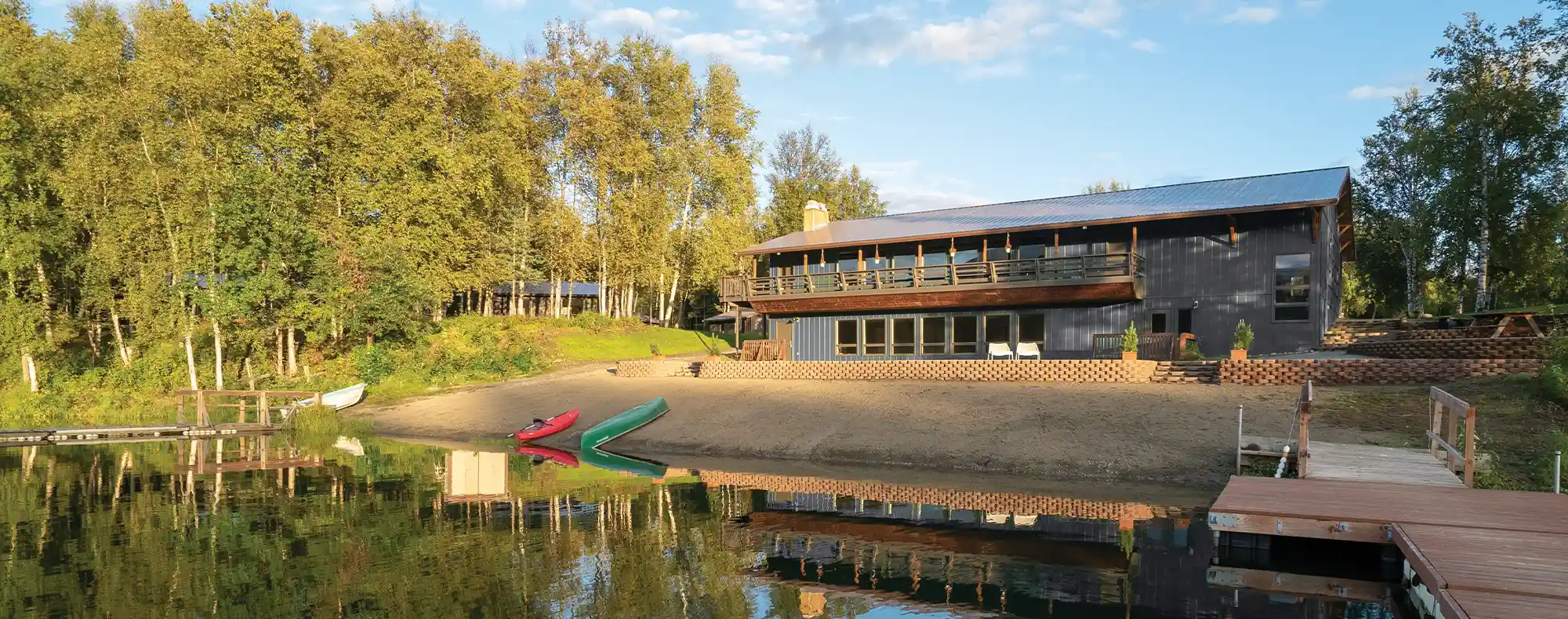 A modern lakeside lodge with a wooden balcony sitting along a sandy beach with canoes, wooden docks, and surrounding trees reflected in calm water.