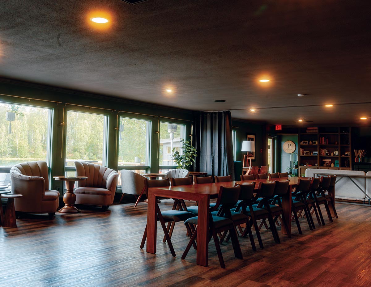 A warm lodge interior featuring a long wooden dining table surrounded by chairs, with plush armchairs arranged along large windows overlooking greenery.