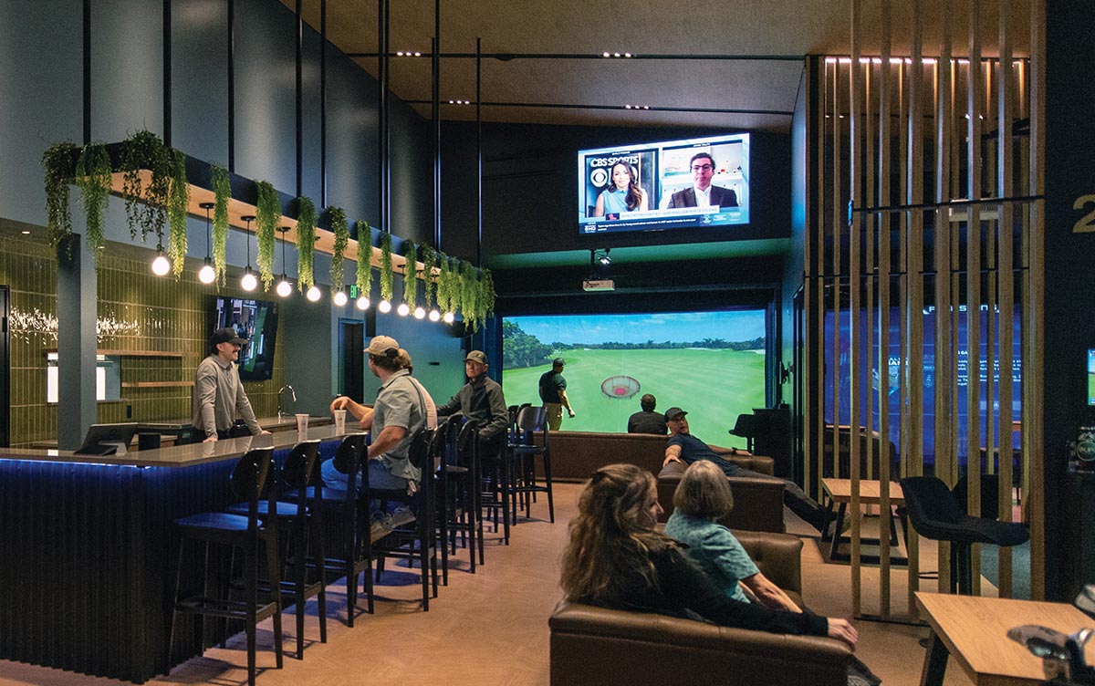 A modern indoor golf simulator lounge featuring a sleek bar with hanging lights, lounge seating, and large virtual golf screens.