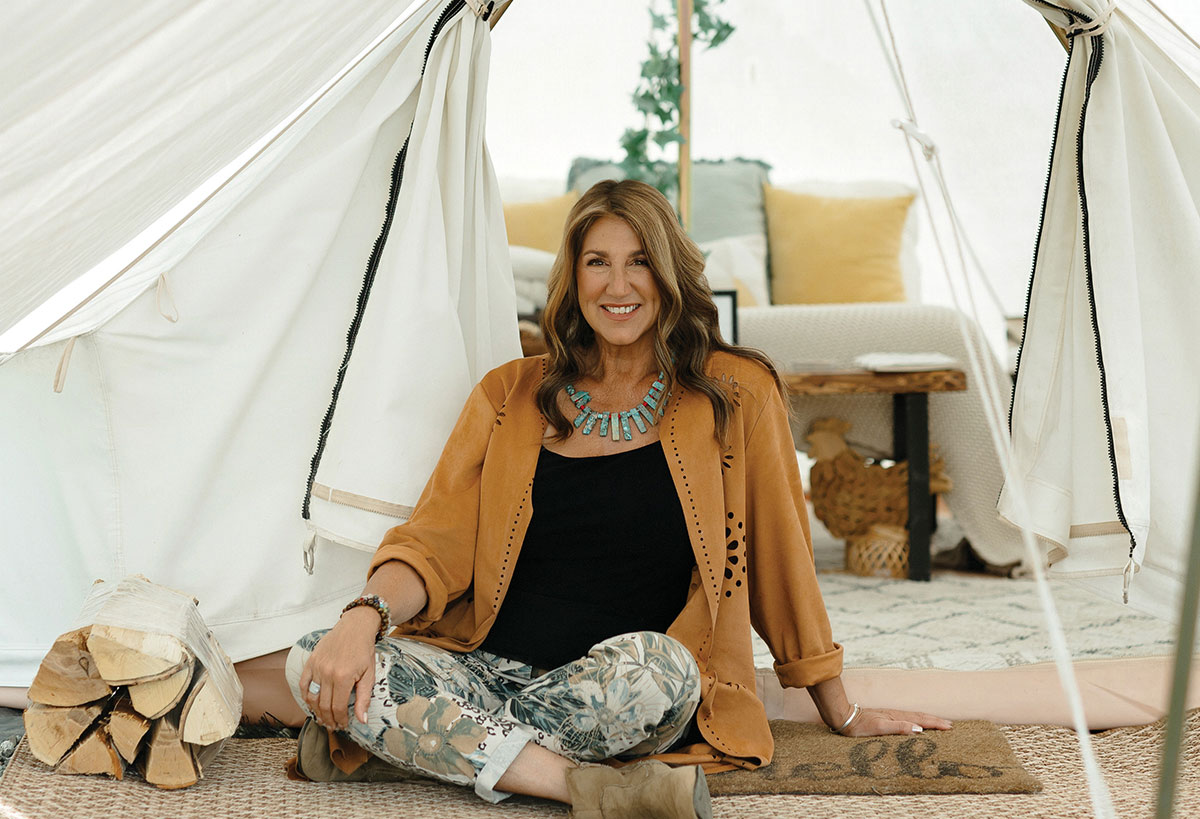 A smiling woman wearing a tan jacket, patterned pants, and a turquoise necklace, sitting cross-legged at the opening of a cozy canvas glamping tent.