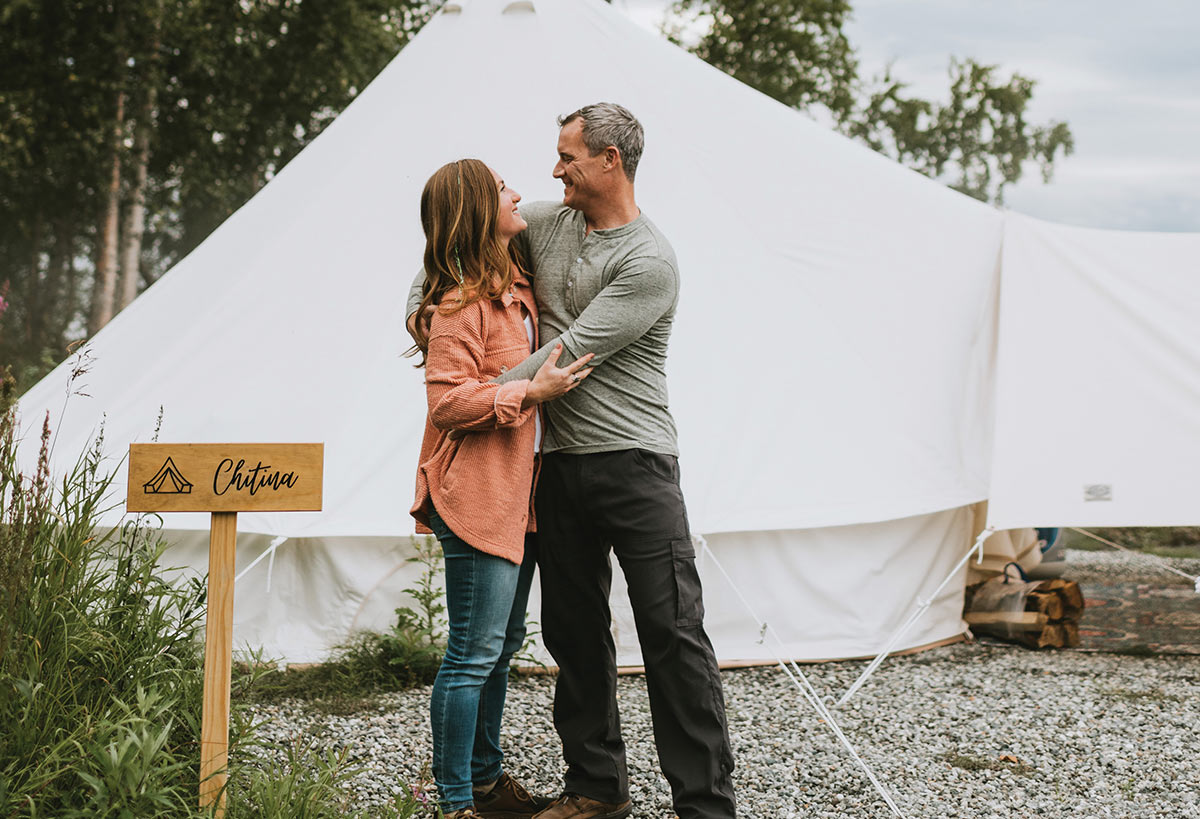 A smiling couple standing in an embrace in front of a large white glamping tent, with a wooden sign reading "Chitina" in the foreground.