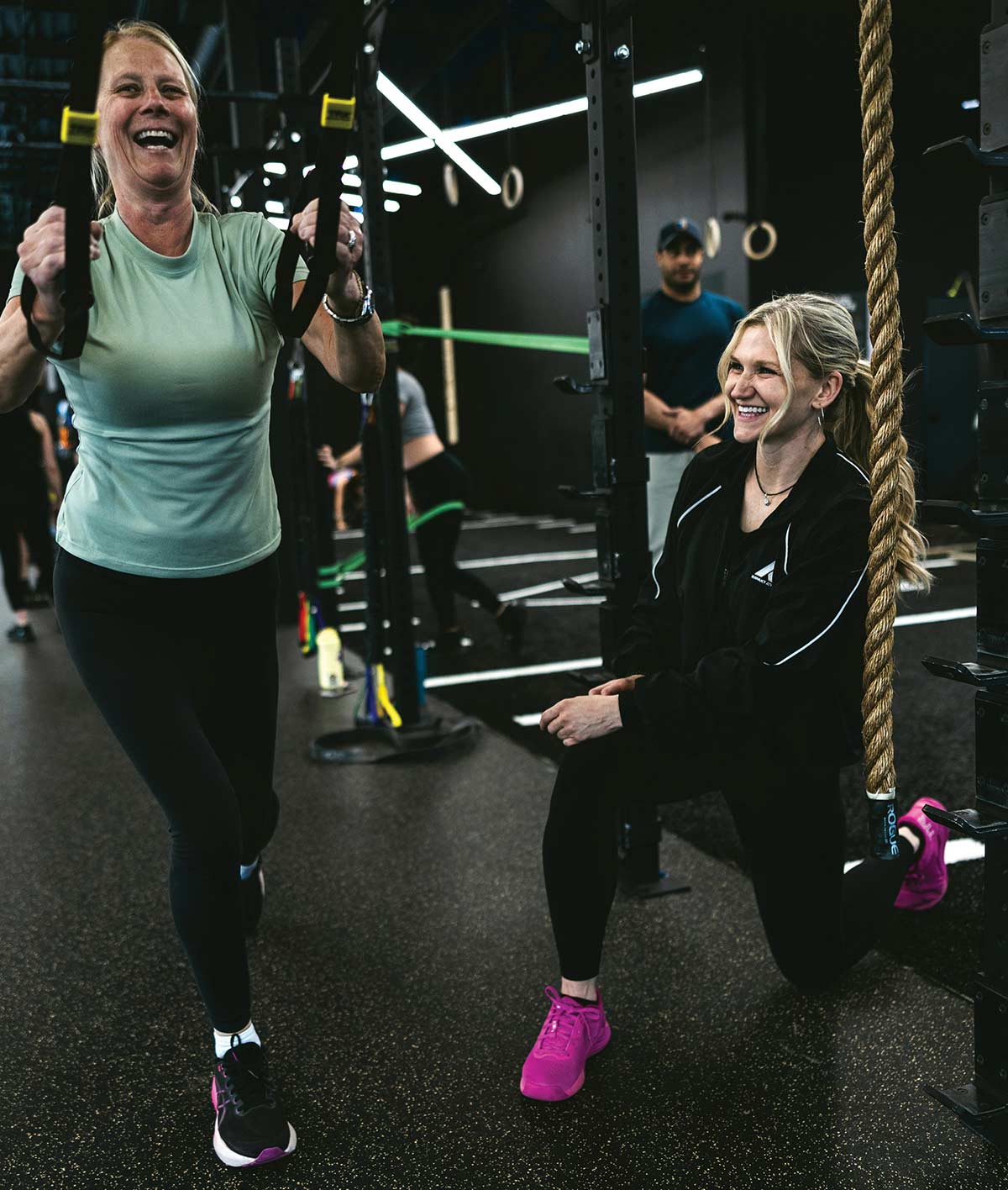A woman laughing while exercising with suspension trainers at a gym, guided by a smiling female trainer kneeling nearby.