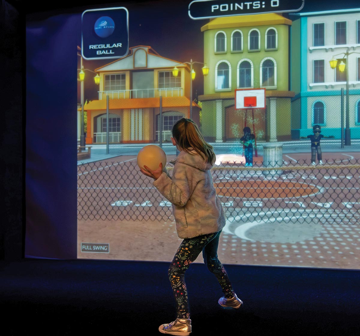 A young girl holding a ball, preparing to shoot at an interactive virtual basketball game projected on a large sports simulator screen.