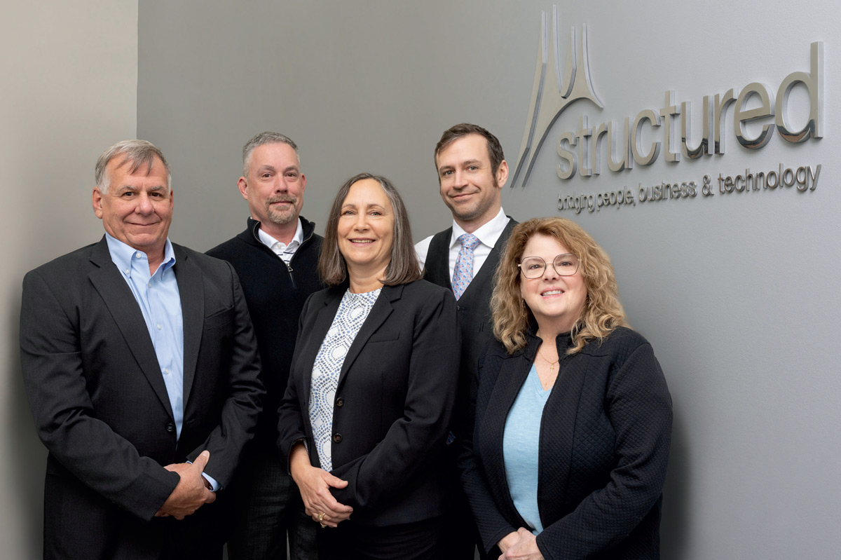 Five business professionals standing in front of a wall with a silver Structured logo.