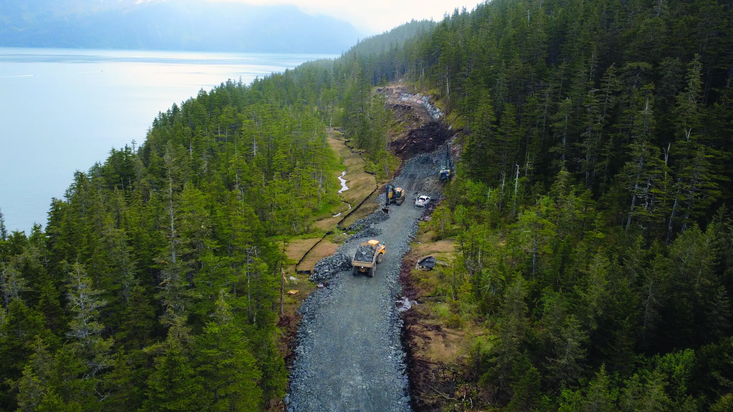 Aerial view of construction vehicles building a gravel access road through forested terrain beside Passage Canal in Whittier.