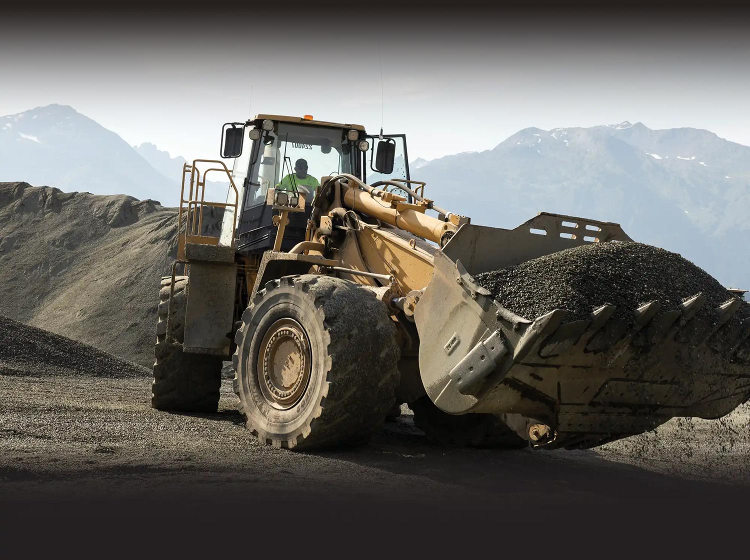 Yellow front loader carrying a heavy bucket full of gravel at an outdoor construction site with mountains in the background.