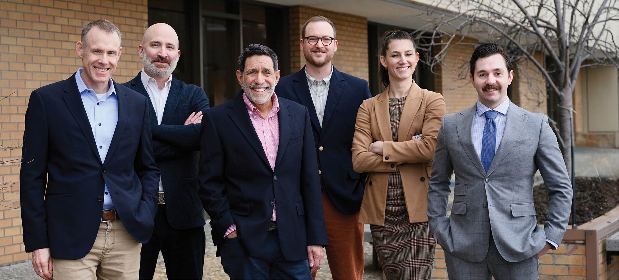 A group of six smartly dressed professionals poses together outdoors in front of a brick building.