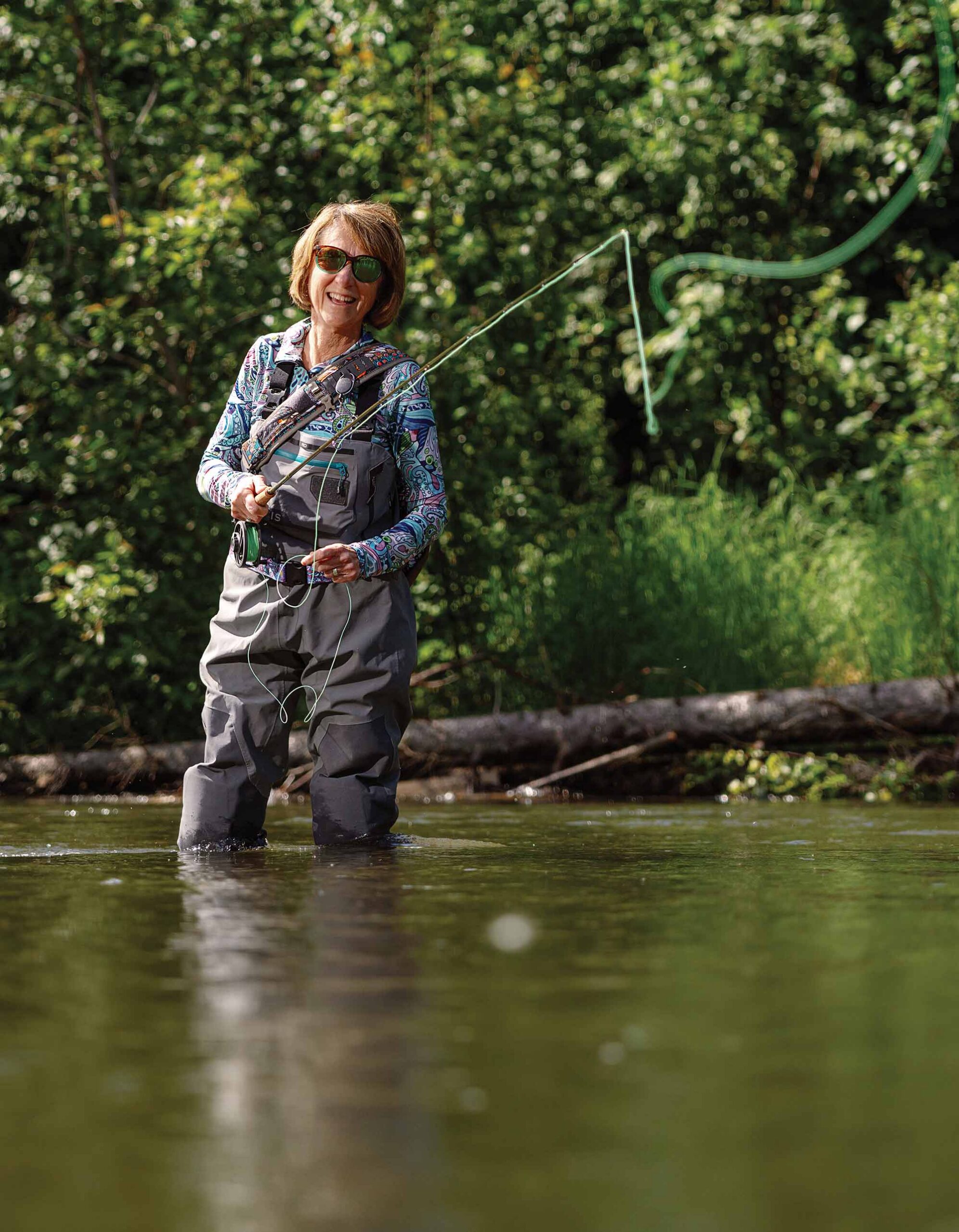 A smiling woman standing in a river casts a fly fishing line.
