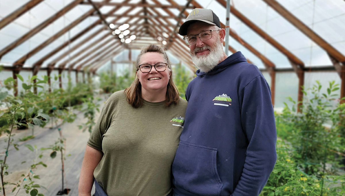 A smiling couple standing together inside a greenhouse filled with potted plants.