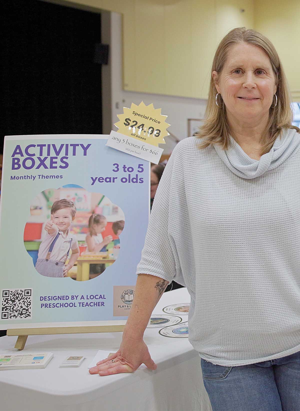 A woman standing next to a display board for "Activity Boxes" designed for 3 to 5 year olds.