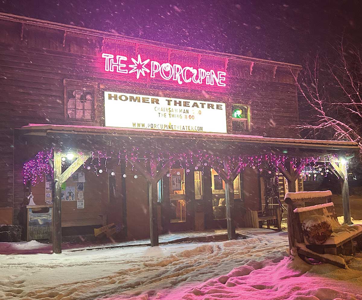 The snowy exterior of "The Porcupine" Homer Theatre illuminated by pink neon lights at night.