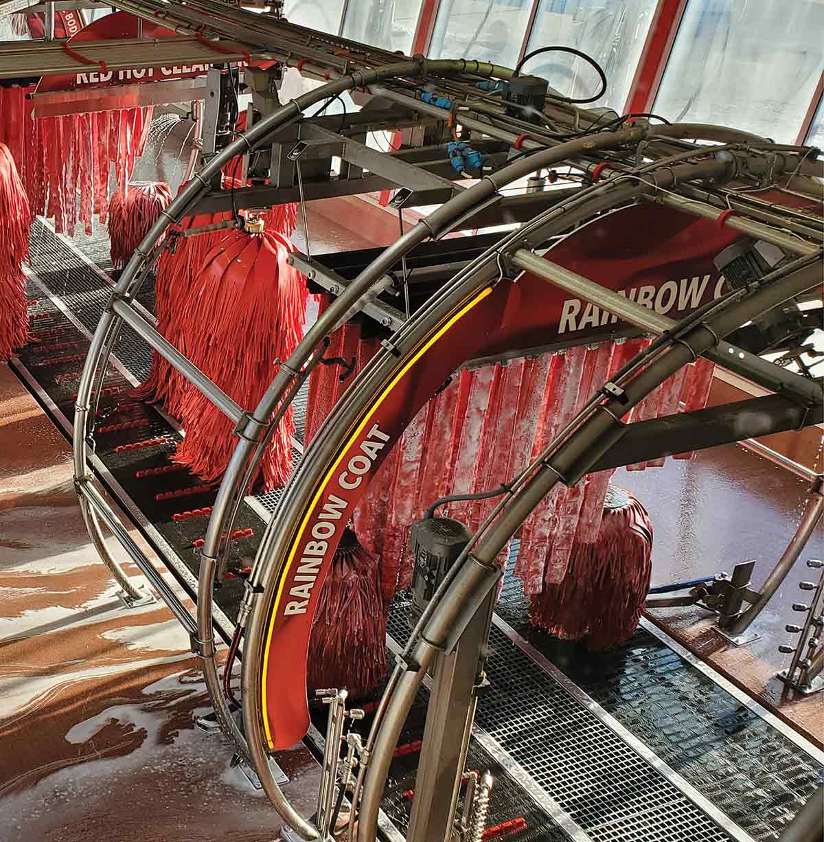 An interior view of an automated car wash tunnel featuring large red brushes and "RAINBOW COAT" spray arches.