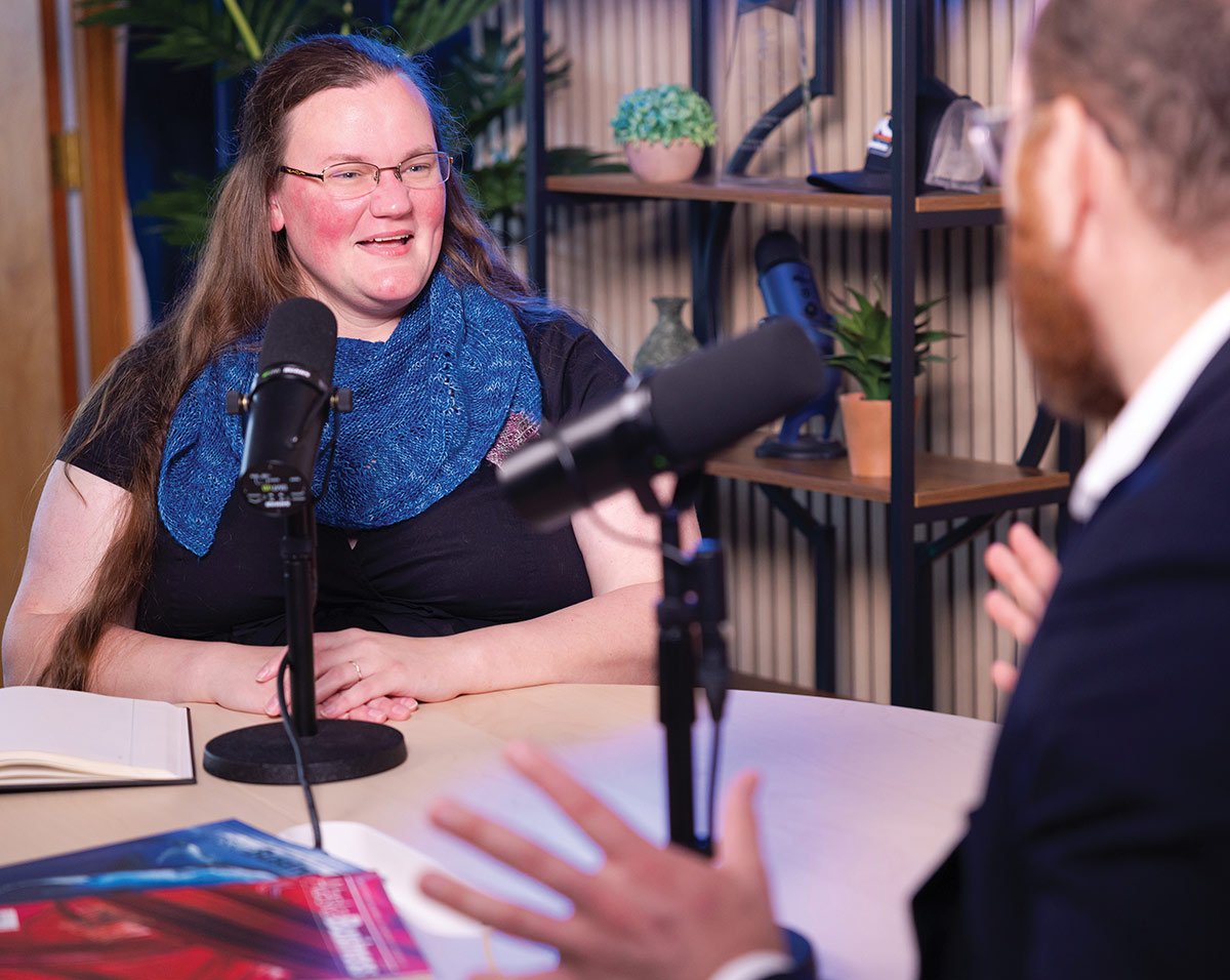 Woman wearing glasses and a blue scarf smiling during a studio podcast interview.