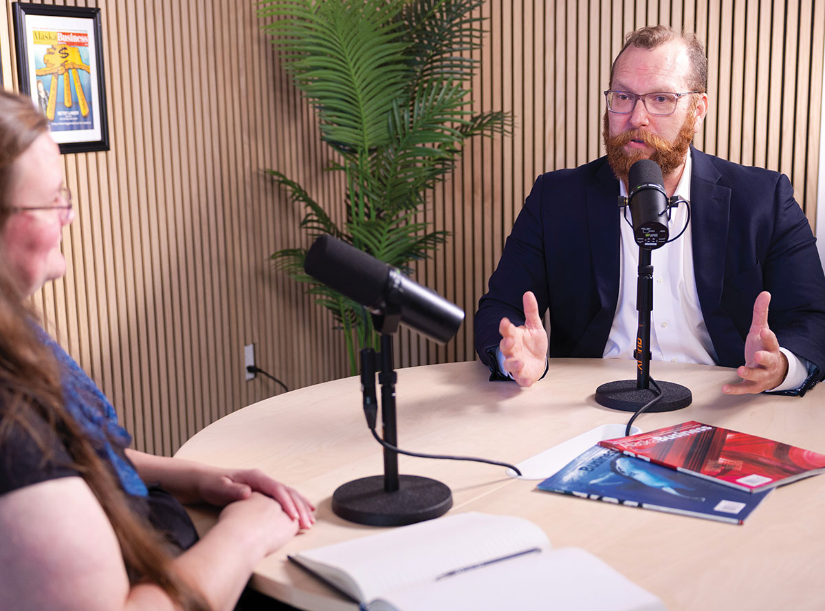 Man speaking into a studio microphone while hosting a podcast seated at a table.