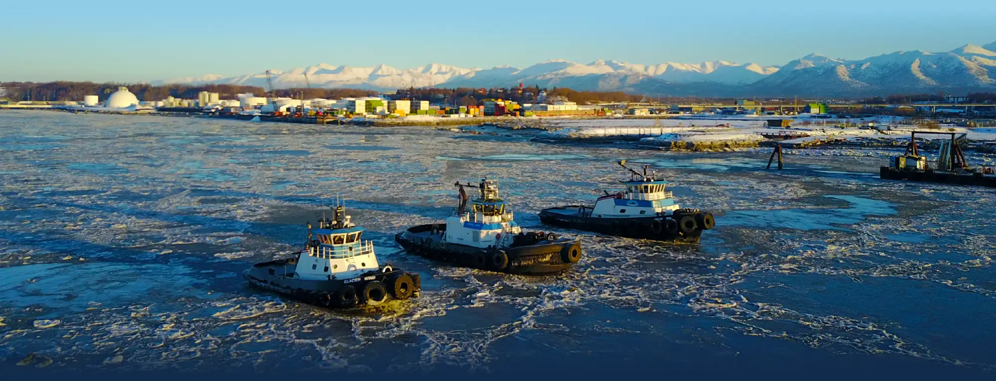 Three tugboats navigating through icy harbor waters, with an industrial port and snow-covered mountains in the background.
