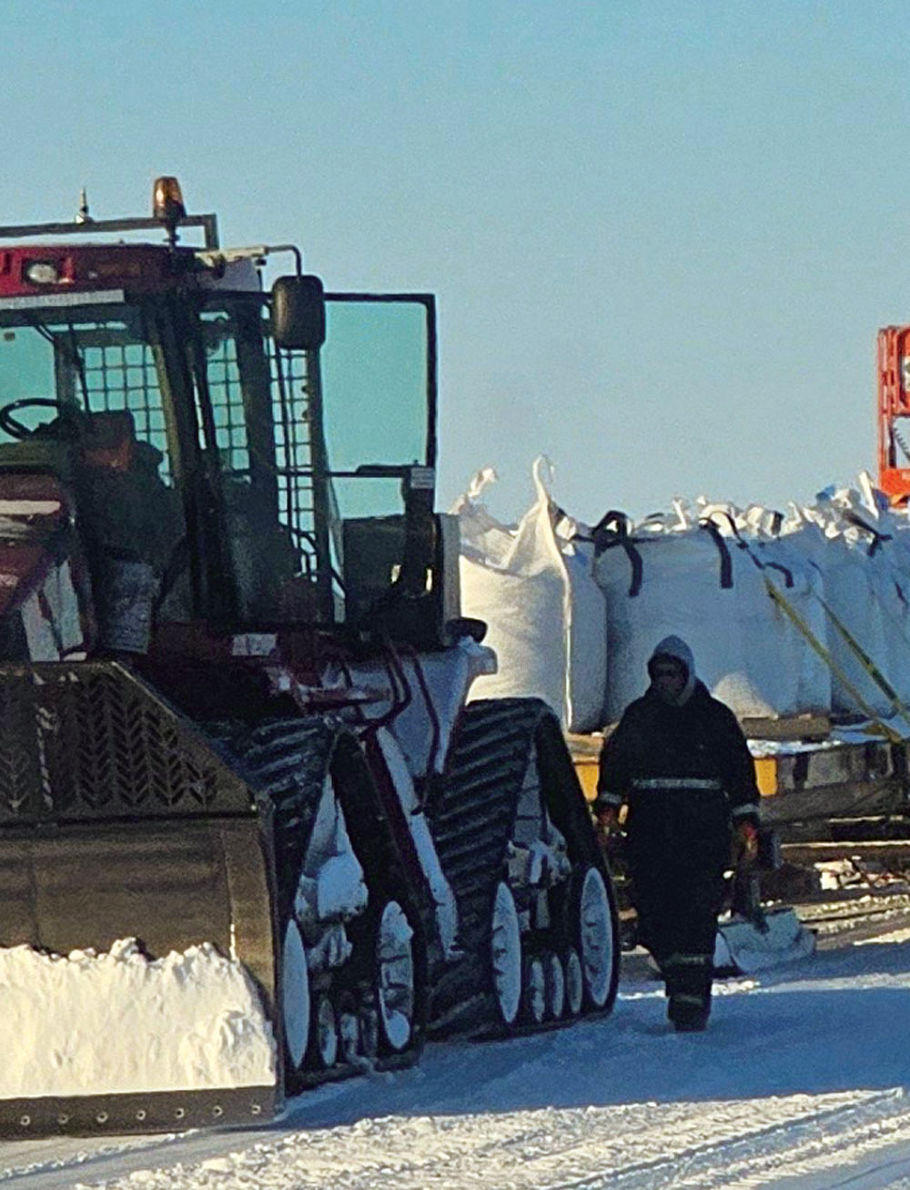 A worker in winter gear walking next to a large tracked snow vehicle towing cargo bags in snowy terrain.