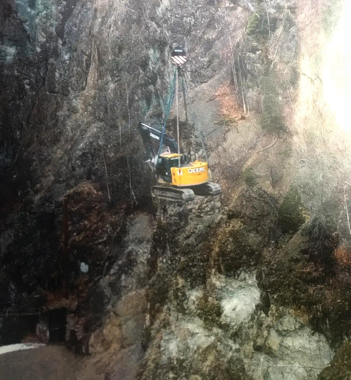 A yellow John Deere excavator hoisted by crane cables along a steep, rocky cliff side.