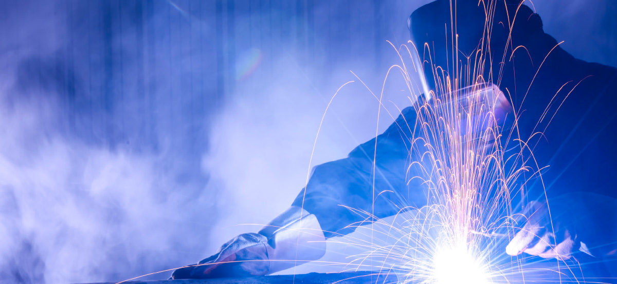 Welder in a protective mask and heavy gloves working as bright sparks spray across a smoky blue background.