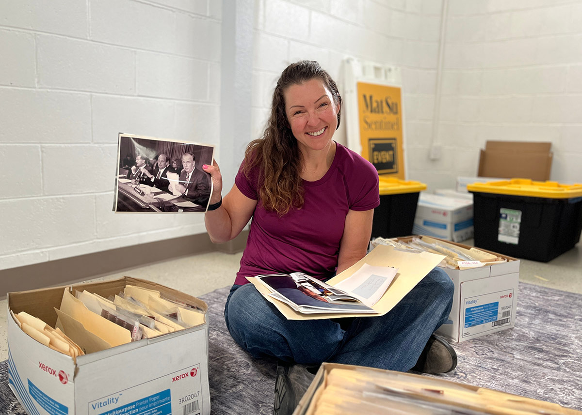 A smiling woman sits on the floor holding up a vintage black-and-white photograph, surrounded by storage boxes filled with archived files and folders.
