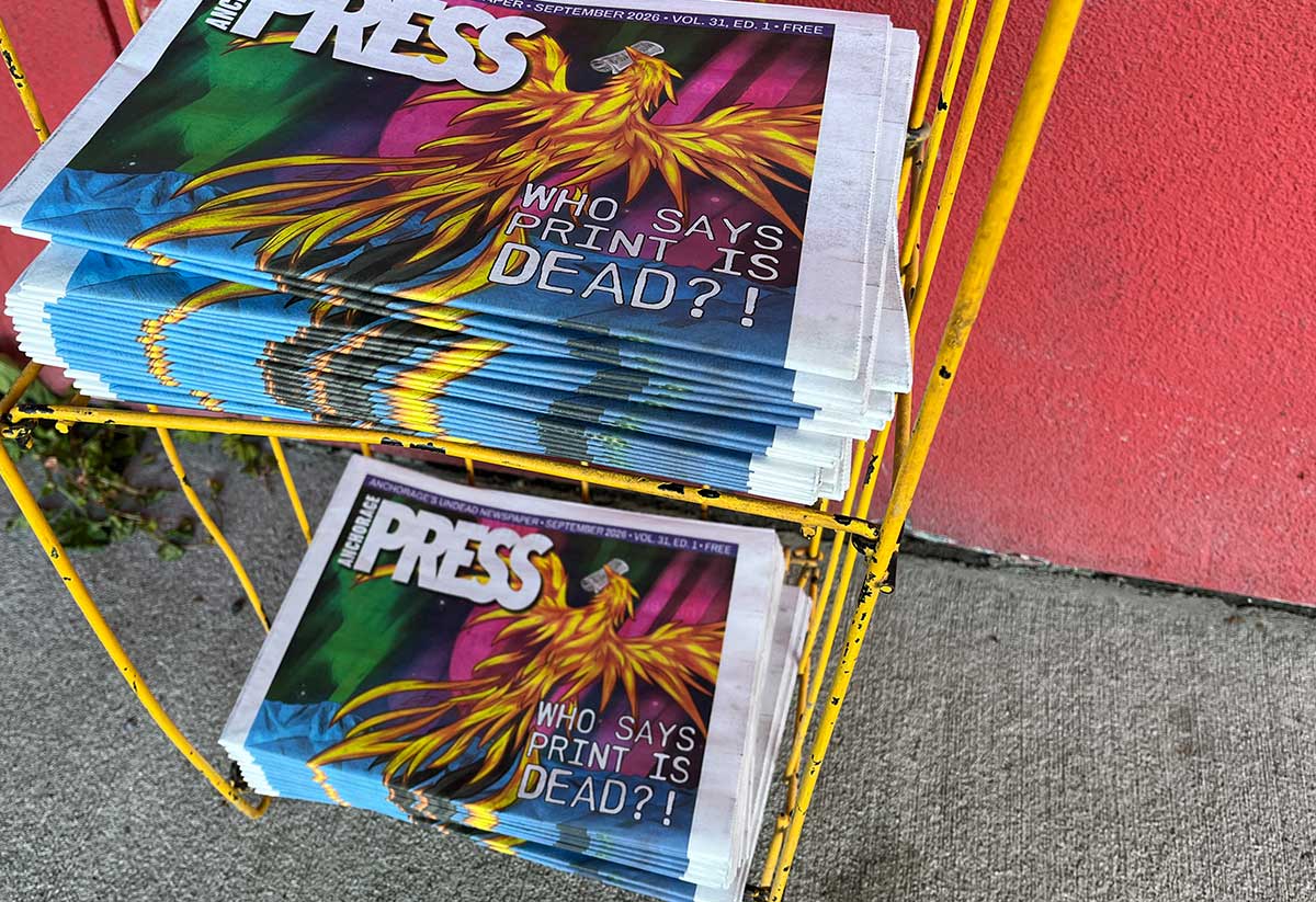Stacks of Anchorage Press newspapers sitting in a yellow wire rack, featuring colorful phoenix cover art and the headline "WHO SAYS PRINT IS DEAD?!".